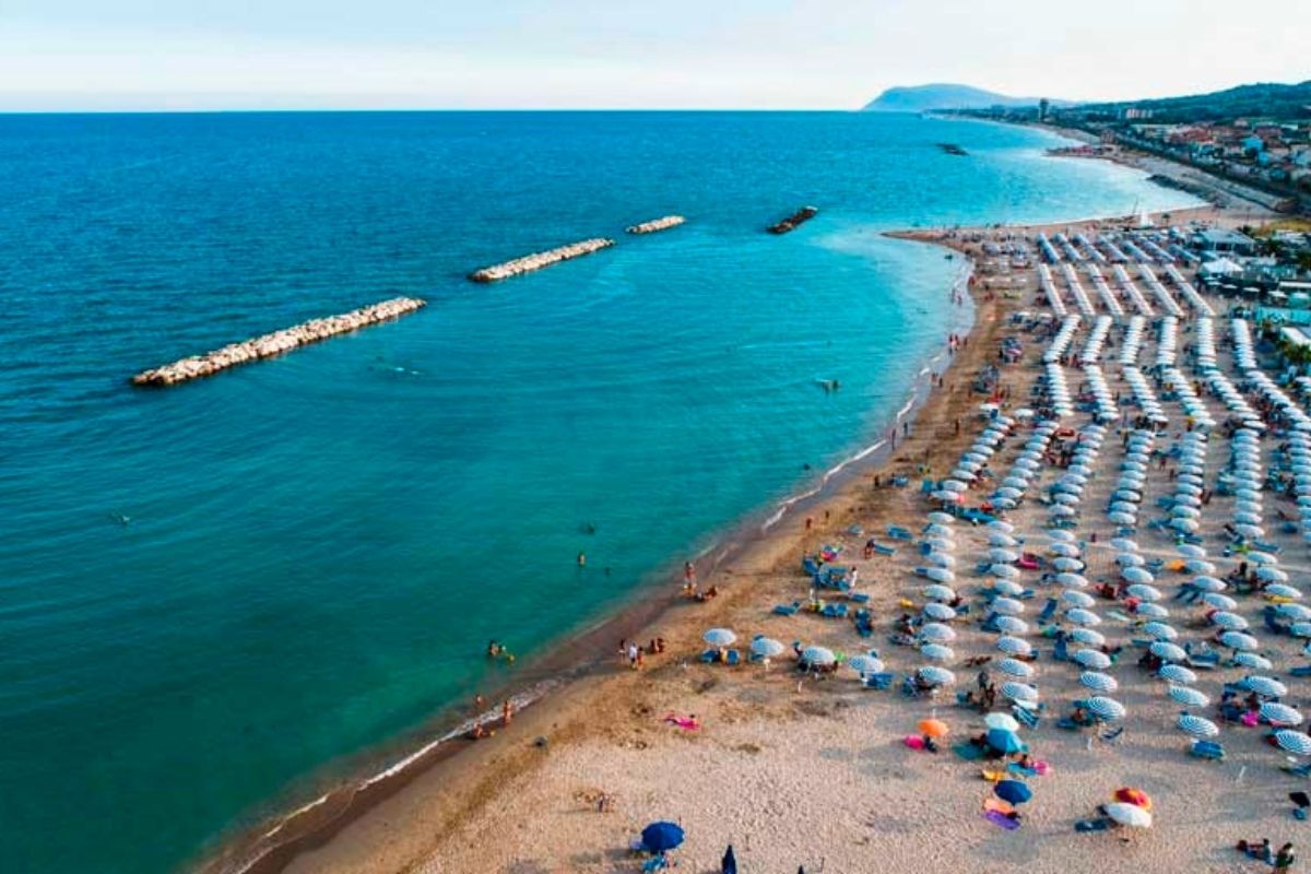 Vista panoramica della spiaggia di Civitanova Marche con persone che si godono il sole, include il porto turistico.